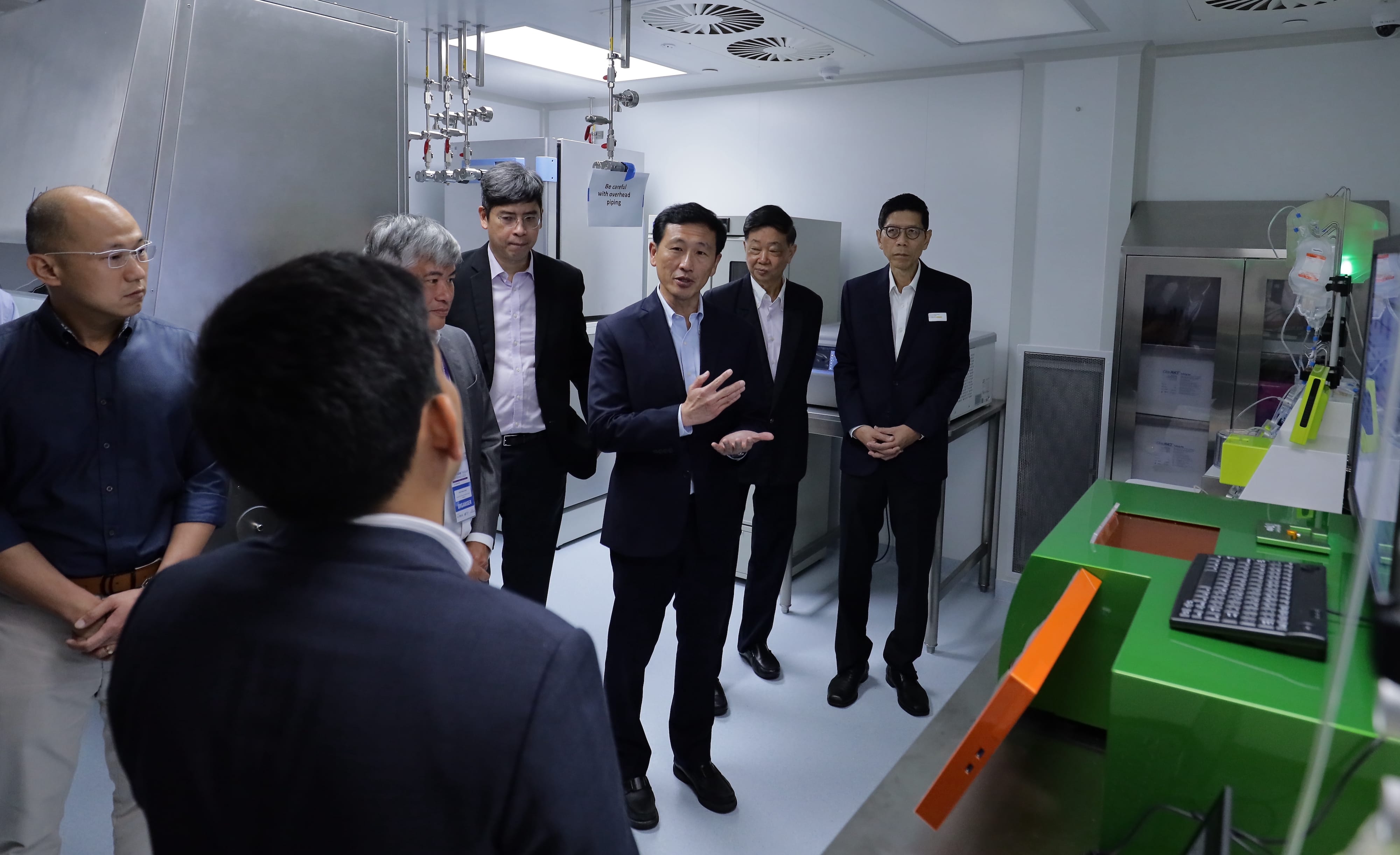 Six men in business attire stand in a lab space near a green machine, one gesturing while speaking.